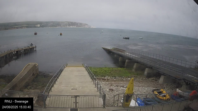 A view of a harbor on a cloudy day, featuring a concrete pier and a ramp leading into the water. Several boats are anchored in the sea, and white cliffs are visible in the distance across the water. There is some green moss or algae on the shoreline near the ramp, and a yellow safety sign with a yellow kayak and orange life jackets are visible in the foreground. The image was captured on June 24, 2025, at 15:00.