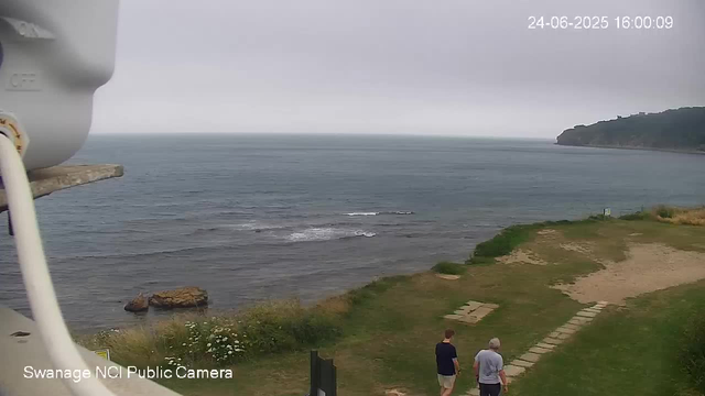 A view of a coastal scene with the ocean and a distant headland under a cloudy sky. Two people are walking along a stone pathway on a grassy area near the shore. A large rock is visible near the water, and there are some white flowers and vegetation along the shoreline. The image is from a webcam labeled "Swanage NCI Public Camera," with a timestamp showing "24-06-2025 16:00:09."