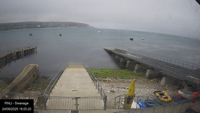 A view of a cloudy day at the harbor showing a concrete pier extending into the water, with a ramp leading down to the shore. Several boats are floating in the calm sea, and distant cliffs can be seen on the horizon. There are small yellow boats or equipment stored on the shoreline, and a yellow caution sign is visible near the bottom right corner. The image is timestamped "24/06/2025 16:00:20" from RNLI station in Swanage.