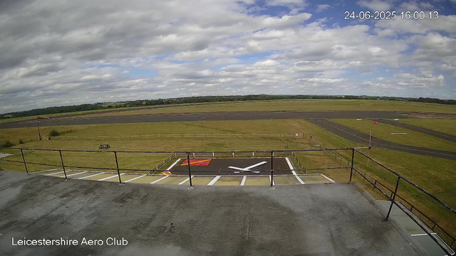 A view from a webcam at Leicesterhire Aero Club showing an empty tarmac area and runway under a partly cloudy sky. The photo captures the airport ground and surrounding fields with a distant horizon and some scattered clouds overhead. The timestamp indicates the date and time as June 24, 2025, at 16:00:13.