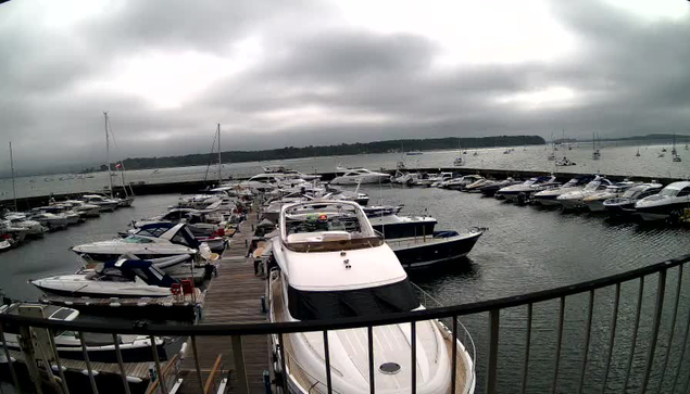 A marina filled with numerous boats and yachts docked along wooden piers, with a cloudy sky overhead and calm water surrounding the vessels.
