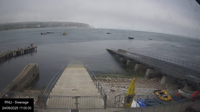 A view of a small harbor with a concrete dock extending into calm water. There are two slipways or ramps leading into the water, one of which appears to be partially submerged. In the foreground, there are yellow safety equipment and a small yellow boat inside a fenced area. Several boats are anchored in the distance on the water, and a foggy, overcast sky hovers over the scene. A cliff or headland is visible in the background across the water.