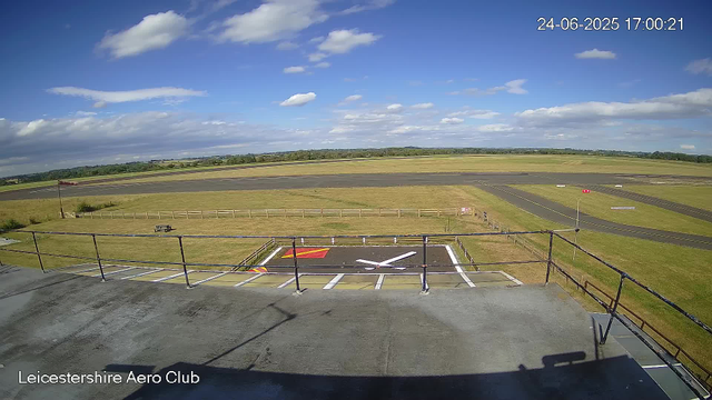 A view from a webcam at Leicesterhire Aero Club showing a bright day with a partly cloudy sky. In the foreground, there is a small helipad with a white helicopter symbol painted on it. Surrounding the helipad is a wooden fence and railing. Beyond the helipad, there is a grassy area with a picnic table and a flagpole with a flag flying. In the distance, there is an airstrip with taxiways and runway markings, set in a rural landscape with open fields and trees under a sky filled with scattered clouds. The timestamp in the upper right corner shows the date as 24-06-2025 and the time as 17:00:21.