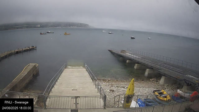 A view of a harbor on a cloudy day with calm water, several boats anchored in the distance, and a rugged coastline in the background. In the foreground, there are piers and safety equipment, including a yellow life jacket and a small boat, all enclosed by a metal fence. The timestamp indicates the date as June 24, 2025, at 6:00 PM.