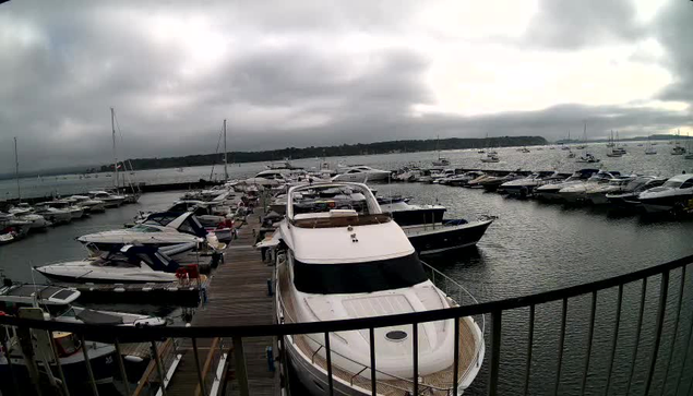 A marina filled with numerous boats and yachts docked along a wooden pier. The water appears calm with boats moored on both sides. The sky is overcast with dark, heavy clouds, and there is a distant shoreline visible on the horizon.