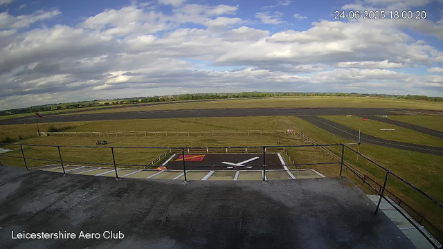 A view from a webcam at Leicesterhire Aero Club showing a runway with a large black and white helicopter tail rotor logo on a paved area. Beyond the runway, there are open grassy fields under a partly cloudy sky with scattered white and gray clouds. The date and time displayed in the upper right corner are 24-06-2025, 18:00:20. The bottom left corner has the text "Leicestershire Aero Club."