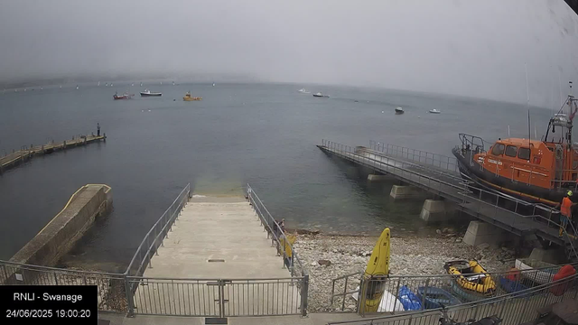 A view of a harbor with boats anchored in the water on a cloudy day. There is a pier extending into the water with a person standing at the end. On the right side, an orange rescue boat is docked next to a ramp. In the foreground, there are yellow and black water rescue equipment and safety signs. The water appears calm with a foggy horizon. The timestamp indicates the image was taken on June 24, 2025, at 7:00 PM.