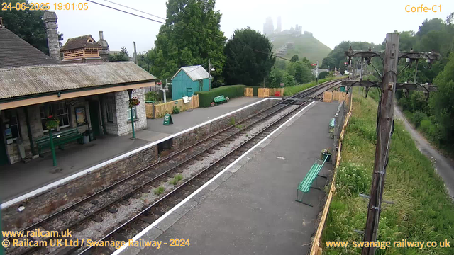 A small train station platform is shown with a stone building on the left, decorated with flower baskets and a green bench. There are two green benches along the platform, with a small teal shed and wooden fencing nearby. On the right side, there are wooden utility poles with wires extending above. The railway tracks run through the center, with some weeds growing between the rails. In the background, lush green trees and hilly terrain are visible, partially obscured by mist or fog, with faint outlines of city buildings on the hilltop. The scene appears to be early evening, based on the timestamp.