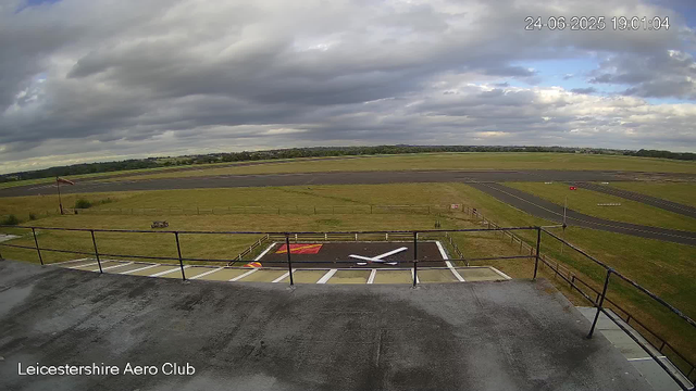 A view from a webcam at the Leicestershire Aero Club showing an empty tarmac area with a large painted white "X" and a red- and yellow-marked section. Beyond the tarmac, there are grassy fields, a runway, and a cloudy sky overhead. The timestamp indicates the date and time as June 24, 2025, at 7:01 PM.