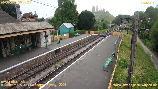 A small train station platform with a stone building on the left, featuring a pitched roof and flower baskets hanging outside. There are several benches along the platform, with a green bench in the center. A light blue shed is situated behind the benches, surrounded by a yellow wooden fence. A "Way Out" sign is placed near the shed. To the right, a set of train tracks runs parallel to the platform, which is bordered by a weathered wooden fence and a row of green benches. Overhead power lines are supported by tall wooden poles. In the background, lush green trees and hills lead up to a hilltop castle with multiple towers and ruins. The image has a timestamp of "24-06-2025 20:00:23" in the top left corner and the text "Corfe-C1" in the top right corner, with website credits at the bottom.