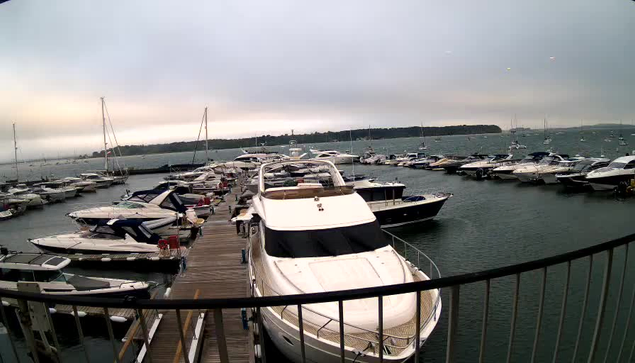 A marina filled with various boats and yachts docked along a wooden pier, with a cloudy sky overhead and a distant shoreline visible in the background.