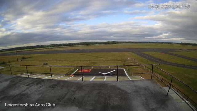 A view from a webcam at Leicestershire Aero Club shows an empty helicopter landing pad marked with a large white "X" and a red rectangle. In the background, there is a grassy area with a small flagpole and a windsock. Beyond the grassy area, there is a paved runway with white lines and markings. The scene is under a mostly cloudy sky with patches of blue, and the timestamp indicates the date and time as June 24, 2025, at 8:00 PM. The landscape is flat with distant trees on the horizon.