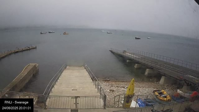 A view of a harbor with a concrete pier extending into the water, surrounded by metal railings. There are small boats anchored in the water in the distance. The sky is overcast, creating a gray and misty atmosphere. A yellow kayak and life jackets are stored near the shoreline, along with some other supplies. The timestamp in the bottom left corner reads "24/06/2025 20:01:01."