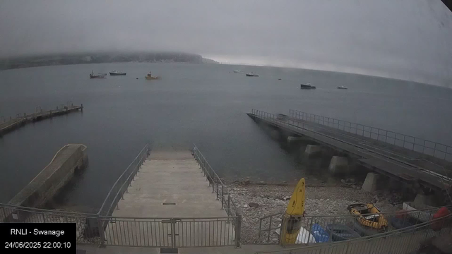 A view of a marina with several boats floating on calm water under an overcast sky. In the foreground, a concrete pier extends into the water with a railing along its edges. To the right, a larger structure extends over the water, supported by concrete pillars. At the bottom right, there is yellow equipment and some other vehicles parked behind a fence. The background shows a foggy shoreline with hills or cliffs partially obscured by mist. The timestamp indicates the image was captured on June 24, 2025, at 22:00:10.
