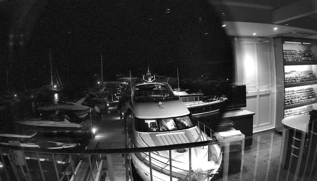 A black-and-white photo taken from a webcam showing a marina at night. Several boats are docked, including a large yacht in the center and smaller boats to the sides. The scene is inside a building with a glass railing in the foreground, and the interior features display shelves filled with objects on the right side. The darkness of the night contrasts with the illuminated interior and the reflections on the water.