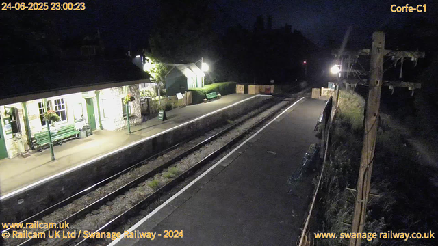 A small train station platform at night, brightly lit with two benches, flower arrangements, and a green hedge. The train tracks run parallel to the platform, which has a "Way Out" sign near the corner. A small building with windows is on the left, and there is a shelter with a roof further down the platform. The surrounding area is dark, with a utility pole and some electrical wires visible on the right side. The time and date displayed at the top left are "24-06-2025 23:00:23".