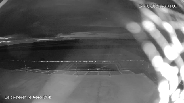 A black-and-white webcam image shows a parking area at night, with a few empty parking spots and a railing in the foreground. The sky is cloudy, and the time and date are displayed in the top right corner as "24-06-2025 23:01:00." The bottom left corner indicates the location as "Leicestershire Aero Club." An out-of-focus object, possibly a chain or rope, is visible on the right side of the image.