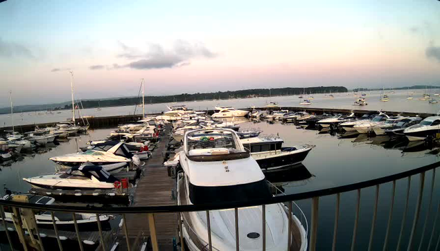 A marina filled with various boats docked along a wooden pier, with calm water reflecting the sky. In the distance, there is a shoreline with trees, under a sky that appears to be during sunset or sunrise with soft light and some clouds.