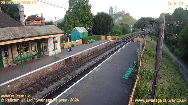 A small train station platform with a stone building to the left, decorated with flower baskets hanging from green poles. There are green benches along the platform, with one near a sign that says "WAY OUT." A green shed is situated behind the benches, with trees and a grassy hill in the background. On the right side, there are railway tracks with wooden fencing along the edge and tall grass beyond. Overhead, there are wooden utility poles holding electrical wires. In the distance, a castle ruins is visible atop a hill, and some signal lights are illuminated. The timestamp on the image shows June 25, 2025, at 5:00 AM.