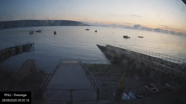 A calm body of water at dawn with several boats scattered across the surface. The sky is mostly clear with soft, light clouds and the horizon shows a distant shoreline and hills. In the foreground, a small dock and a ramp lead down to the water, with some rocky and grassy terrain nearby. The timestamp indicates the photo was taken early in the morning.