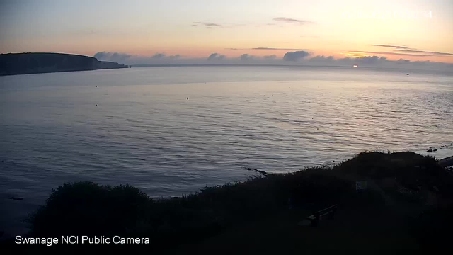 A view of a calm sea at sunset, with the sky painted in soft pastel colors. In the distance, there are some clouds along the horizon. To the left, a dark headland extends into the water. The foreground features dark bushes and a small section of land along the shoreline. The image is from a public webcam in Swansea.