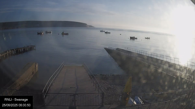 A view of a calm harbor with several boats anchored in the water. There are two wooden piers extending into the harbor, with one in the foreground and the other to the left. The sky is partly cloudy, and the sun is shining brightly from the right side, creating a lens flare effect. In the background, there is a hilly shoreline with cliffs. The timestamp indicates early morning on June 25, 2025.