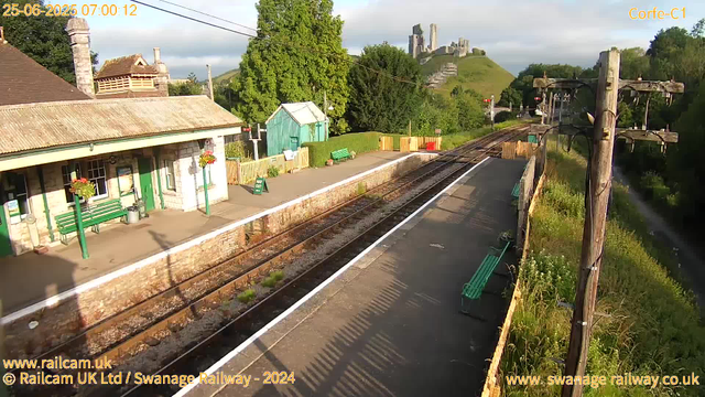 A small train station platform with green benches and a green building is visible in the foreground. There are flower baskets hanging on the building's exterior. The platform is empty, and there are train tracks running alongside it. A wooden fence, some colorful containers, and a small green shelter can be seen on the far side of the platform. In the background, a lush green hill with what appears to be a historic castle or ruins perched on top is visible, with the surrounding area filled with trees and greenery. The sky is partly cloudy with some sunlight illuminating the scene.