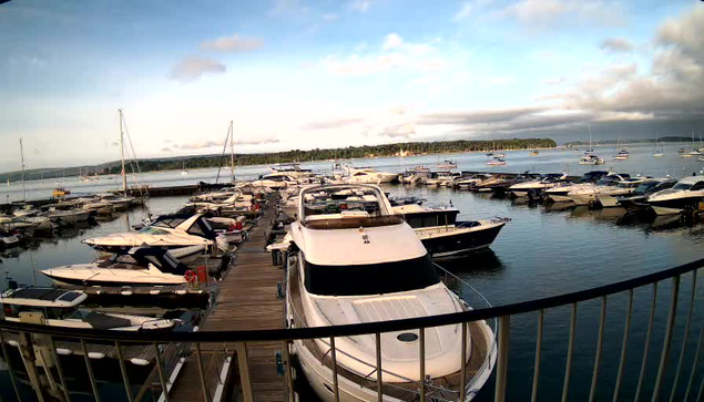 A marina filled with various boats and yachts docked along wooden piers. The water is calm, and the sky above is partly cloudy with patches of blue. In the background, there is a distant shoreline with trees. The image is taken from a viewpoint behind a black metal railing.