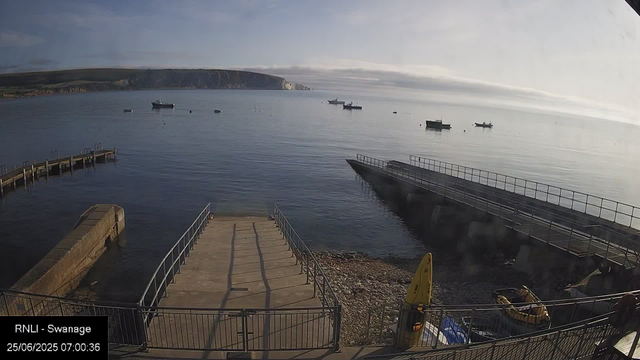A view of a harbor with calm water and several boats floating in the distance. There is a small wooden dock extending into the water on the left side, and a sloped concrete ramp with metal railings leading down to the water in the center. On the right side, there are two small yellow boats, a yellow caution sign, and a person standing near the fence. In the background, a hilly coastline is visible under a partly cloudy sky with the sun shining gently. The timestamp indicates the photo was taken on June 25, 2025, at 7:00 AM.