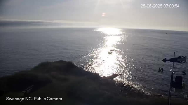 A view of the ocean at sunrise, with the sun reflecting brightly off the water's surface. In the foreground, there is a grassy, slightly elevated land area and a weather vane pointing north and south. The scene appears calm and peaceful with no visible boats or people.