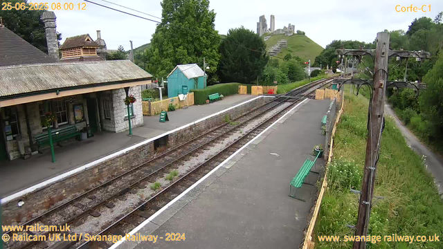 A small train station platform with a stone building to the left, featuring a tiled roof and hanging flower baskets. The platform has green benches and a small blue shed towards the back, surrounded by a yellow fence. There are railway tracks in the center of the image, with vegetation growing between the sleepers. To the right, there is a row of wooden electric poles with wires overhead, and a grassy area with wildflowers and a narrow footpath along the edge of the platform. In the background, a hill with ancient ruins or a castle remains can be seen atop the green landscape. The sky is overcast, and the timestamp shows the date as June 25, 2025, at 8:00 AM.