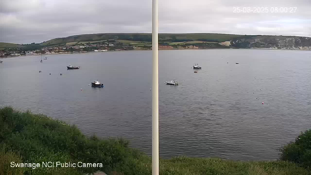 A view of a calm body of water with several boats anchored near the shore. In the background, there are hills with patches of greenery and some buildings along the coastline. The sky is overcast with gray clouds. The timestamp in the top right corner indicates the date and time as 25-06-2025 at 08:00:27. The bottom left corner displays the text "SwaHage NCI Public Camera."