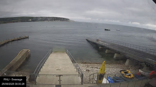 A view of a harbor with a small dock and a pier extending into calm water. Several boats are anchored in the distance under a cloudy sky. There is a yellow safety sign and some equipment near the shoreline, with rocky terrain visible at the edge of the water. In the background, a cliff coastline stretches along the horizon. The timestamp shows "25/06/2025 09:00:28".
