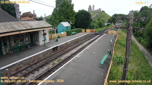 A small train station platform with a stone building on the left, decorated with hanging flower baskets. There are several green benches along the platform, and a sign indicating "WAY OUT." The railway tracks run through the center, with a footpath and greenery on the right side. In the background, there is a small blue shed, a hill with lush green trees, and a castle ruin on top of the hill. An old wooden utility pole with wires extends from the foreground toward the right. The image is marked with the date and time at the top left corner.