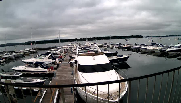 A marina filled with numerous yachts and boats moored alongside a wooden dock under a cloudy sky. The water is calm, and a railing is visible in the foreground.
