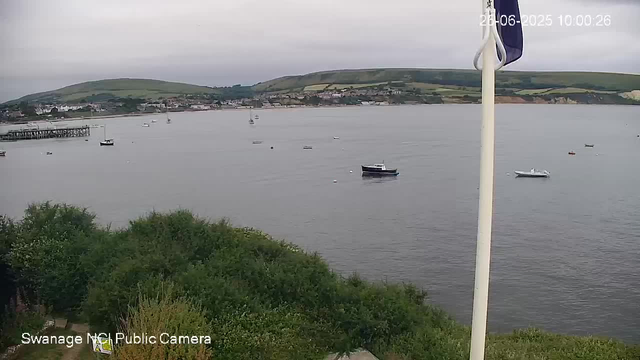 A calm body of water with a few boats floating on its surface, surrounded by green hills in the background. There is a cloudy sky overhead, and some greenery and trees in the foreground. A white flagpole with a flag attached is partially visible on the right side of the image. The timestamp in the top right corner reads "26-06-2023 10:00:26."