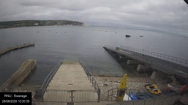 A view of a harbor with a concrete slipway leading into calm water, surrounded by railings. Several small boats are anchored in the water, and some are near the slipway. The sky is overcast with thick clouds, and a distant shoreline with cliffs and green hills is visible across the bay. There are a few birds perched on the railings and boats.