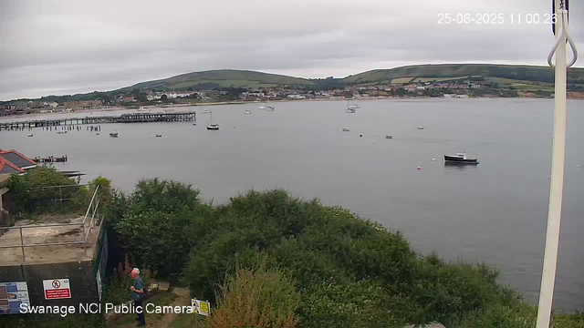 A view of a harbor with calm water and several small boats floating. In the background, there are rolling green hills and a small town visible along the shoreline under a cloudy sky. In the foreground, there is greenery and a person standing near some signs, with a building partially visible on the left. The timestamp on the image is "25-06-2025 11:00:26".