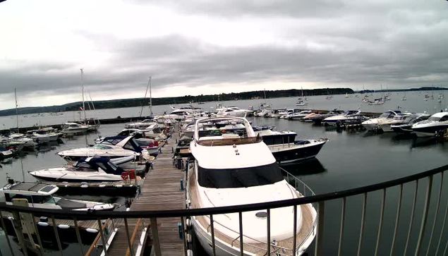 A marina filled with numerous white and dark-colored boats docked along wooden piers. The boats vary in size, with some large yachts and smaller vessels. The scene is under a cloudy sky, and a railing is visible at the bottom of the image, indicating the photo was taken from a vantage point overlooking the marina.