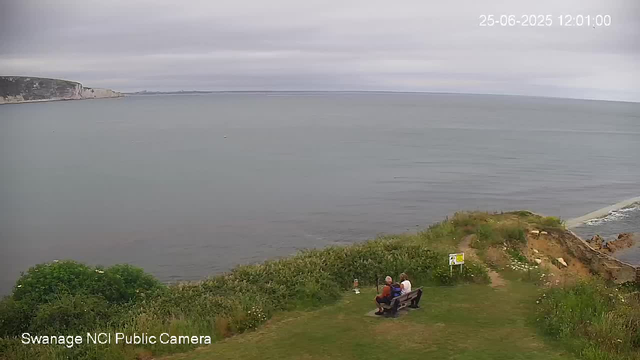A view of a calm sea with overcast skies, a rocky shoreline with green vegetation, and two people sitting on a bench facing the water. The time and date are displayed in the top right corner, and a small sign is near the bench on the right.