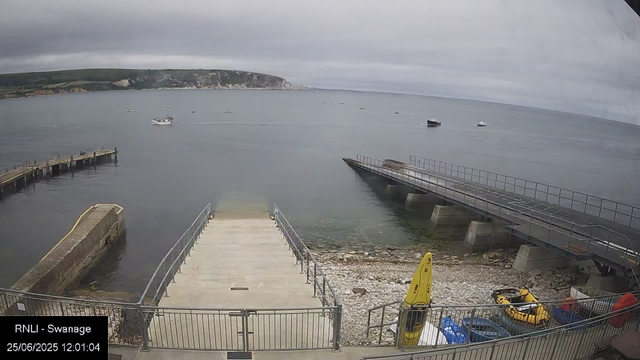 A view of a harbor with a concrete boat ramp leading into calm water, a metal pier extending into the water on the right, and a small pier on the left. Several boats are floating in the water, and there are yellow safety floats and equipment near the shore. The background features a hilly coastline under a cloudy sky. The timestamp indicates it was taken on June 25, 2025, at 12:01 PM.
