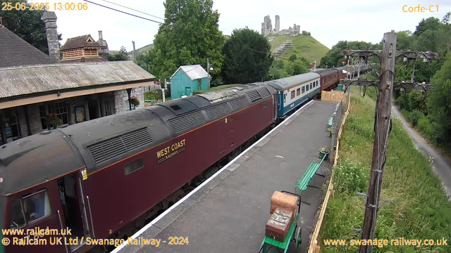 A vintage steam train with maroon and blue carriages is stopped at a small rural station platform. Surrounding the station are green trees, a stone building with a tiled roof, and a small turquoise shed. In the background, there is a grassy hill with a partially ruined castle ruin on top. To the right of the platform, a wooden utility pole with multiple wires and a grassy path runs alongside. The scene appears peaceful, with a partly cloudy sky and lush greenery. The timestamp on the image indicates the date and time as June 25, 2025, at 1:00 PM.