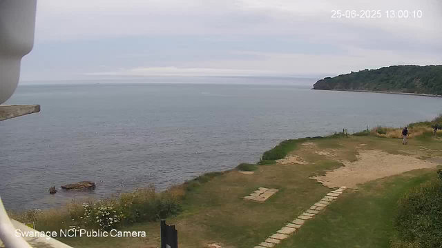 A wide view of a coastal scene with a calm ocean, a small rocky outcrop near the shore, and a distant headland covered in greenery. There is a grassy area with a stone pathway, and a few people walking along the path on the right side of the image. The sky is overcast with grey clouds. The timestamp in the top right corner shows "25-06-2025 13:00:10."