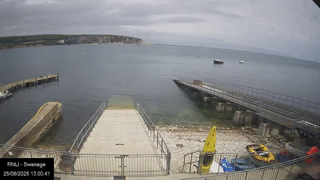 A view of a seaside dock area with a small pier extending into calm waters. There are three small boats anchored nearby and a larger boat further out. The coastline with cliffs is visible in the background. In the foreground, there are yellow and orange equipment and a fenced area with kayaks and other gear. The sky is mostly cloudy.