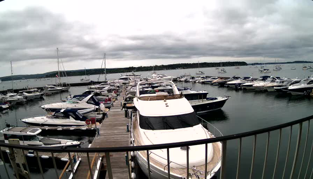 A dock extending into a harbor filled with numerous boats and yachts of various sizes. The sky is overcast with dark, heavy clouds. The foreground features a curved black railing.