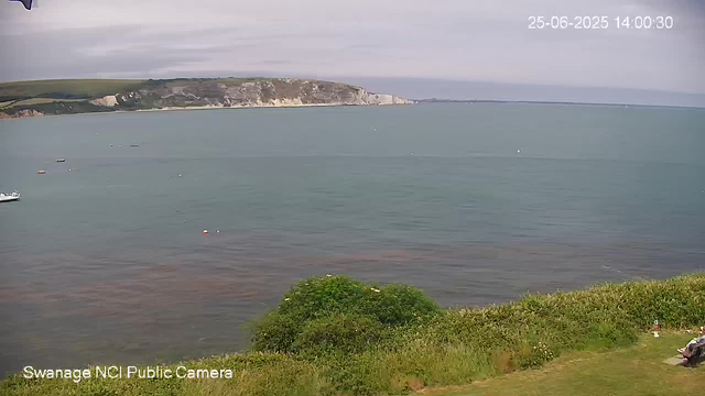 A panoramic view of a coastline with a calm sea and a cliff in the background. The sky is partly cloudy. In the foreground, there is green vegetation with a small bench and a person sitting on it. There are a few boats floating on the water. The date and time are displayed in the top right corner as "25-06-2025 14:00:30".