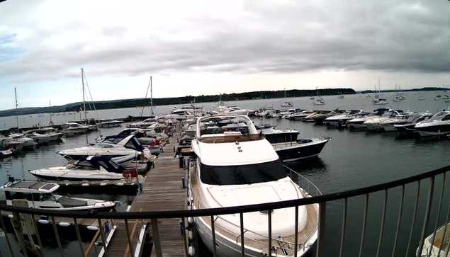 A marina filled with various boats and yachts docked along wooden piers. The water is calm, and there are numerous sailboats with masts visible in the background. The sky is overcast with gray clouds.