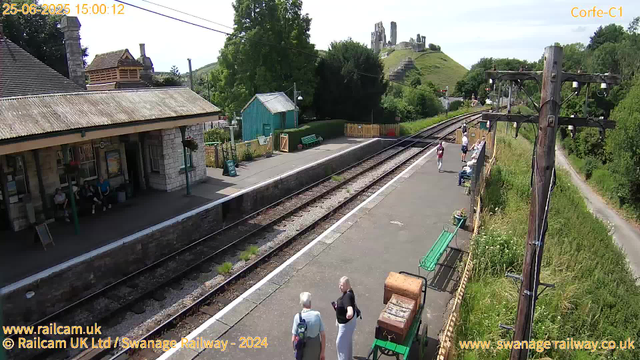 A small train station platform with a stone building on the left, where a few people are sitting. Two people are standing and talking near the bottom center. Several more people are visible further down the platform, some sitting on benches and others walking. There is a green bench and a small wooden cart with flowers on it. Overhead, there are electricity poles with wires, and in the background, there is a hill with ruins or old stone structures on top, with greenery and trees around. The station appears peaceful and sunny.