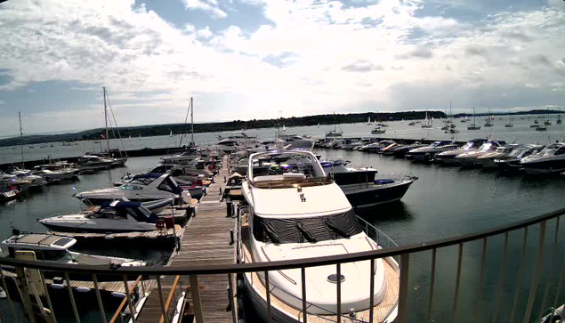 A marina with numerous sailboats and motorboats docked along wooden piers. The sky is partly cloudy, and the water is calm, reflecting the boats. The photo is taken from an elevated position behind a curved metal railing.