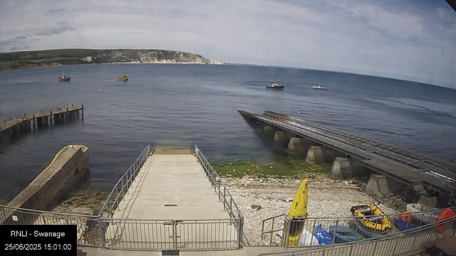 A view of a seaside dock area with a paved platform and metal railings in the foreground. To the right, there are yellow water safety floats and small boats, including an inflatable yellow boat and some water gear. The water shows mild waves, and there are four small boats floating in the distance. A low pier extends into the water on the left, with a background of green, white cliffs under a partly cloudy sky. The bottom left corner displays text indicating the location as "RNLI - Swanage" and the date as "25/06/2025" with the time "15:01:00."
